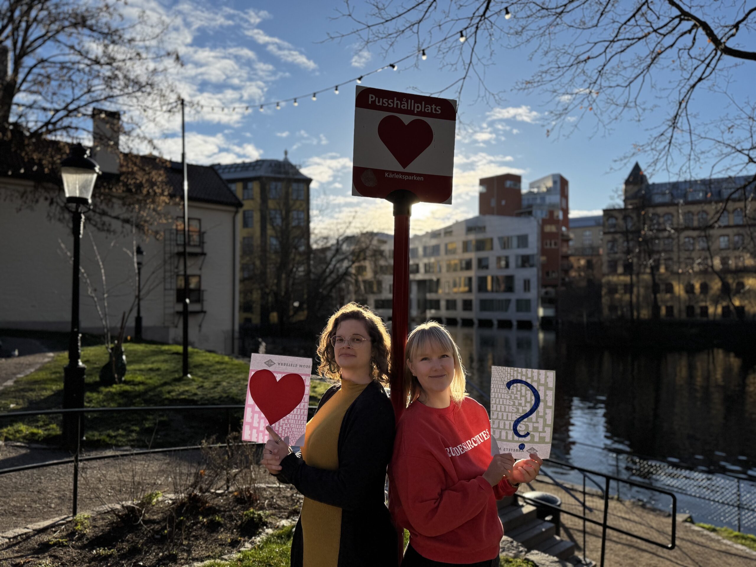 Sofia och Ida står vid en skylt med texten pusshållplats. De håller varsin skylt, en föreställer ett hjärta och en föreställer ett frågetecken. De är utomhus och det är soligt.